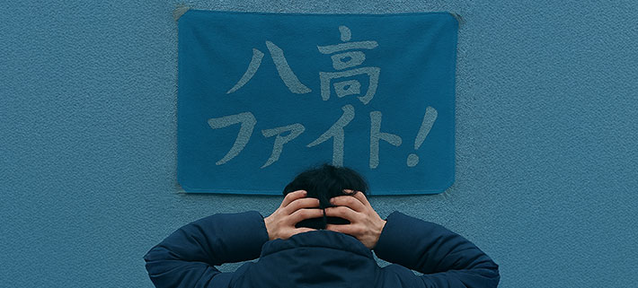 青色の生地の上に「八高ファイト！」という文言を、薄い青色で名入れ印刷した視認性が低いオリジナル応援旗の前で頭を抱えている人の画像