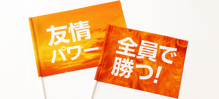 オレンジ色夕暮れの空を思わせる、オレンジのグラデーション生地の中央に、白い太字で大きく「友情パワー」と名入れ印刷された応援旗と、炎を思わせるオレンジ色のグラデーション生地の中央に白い太字で大きく「全員で勝つ！」と名入れ印刷された応援旗が二つ並んでいる画像
