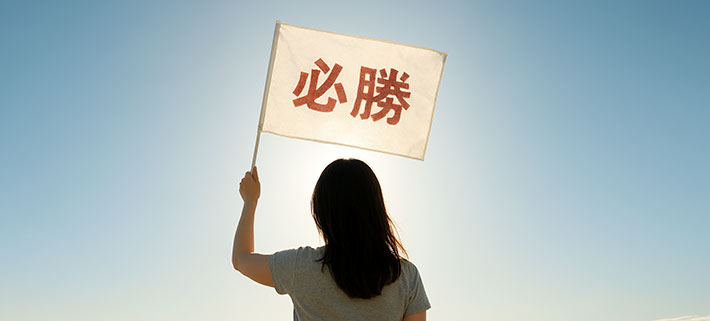 白色の生地に「必勝」という文字を、薄い赤色で名入れ印刷された視認性が低いオリジナル応援旗を頭の上で掲げ、背を向けている女性の画像